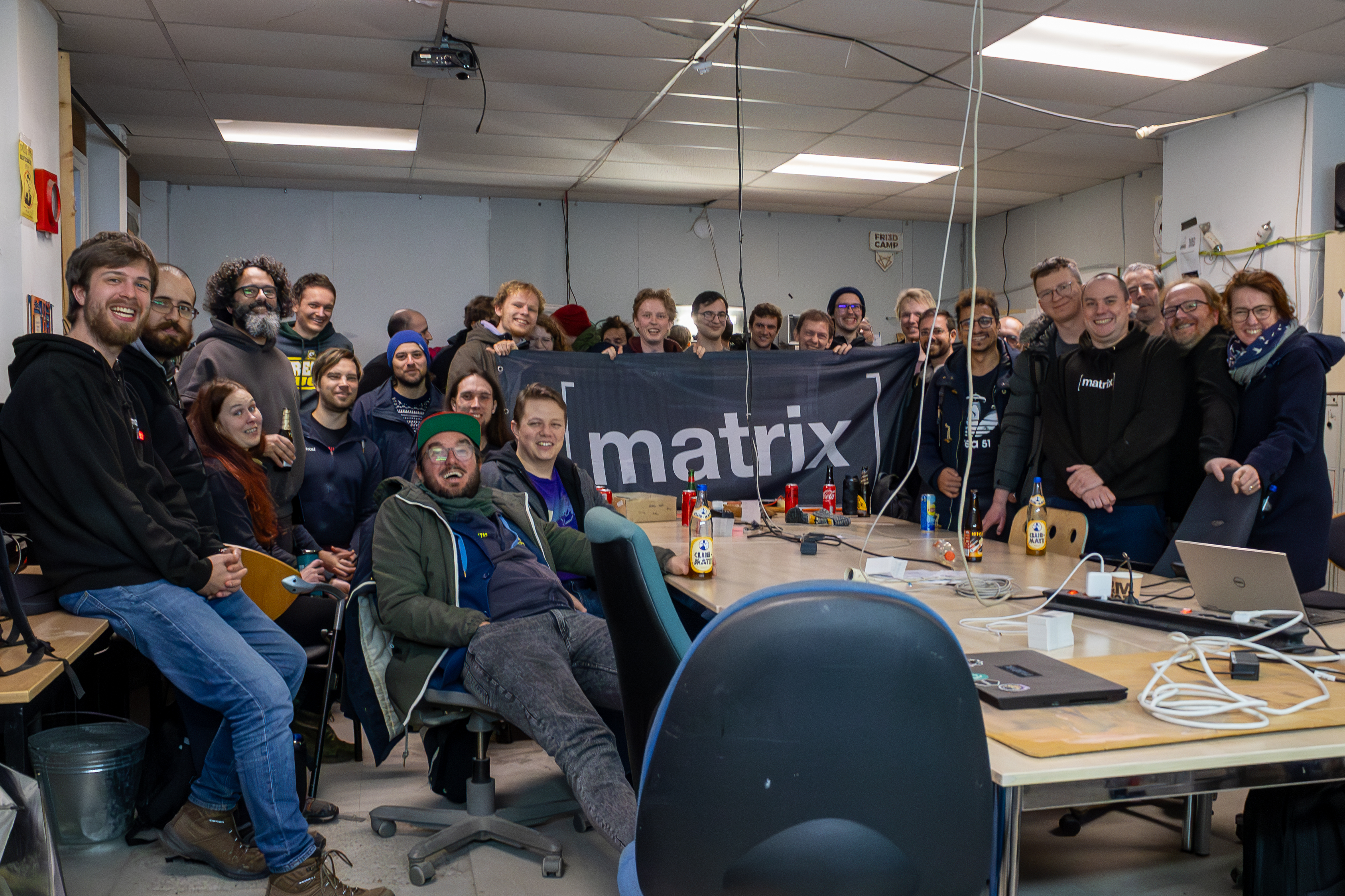A group picture taken at the Matrix Community Meetup in Brussels showing around 30 attendees in a crowded hackerspace room with several Club Mate bottles on the table